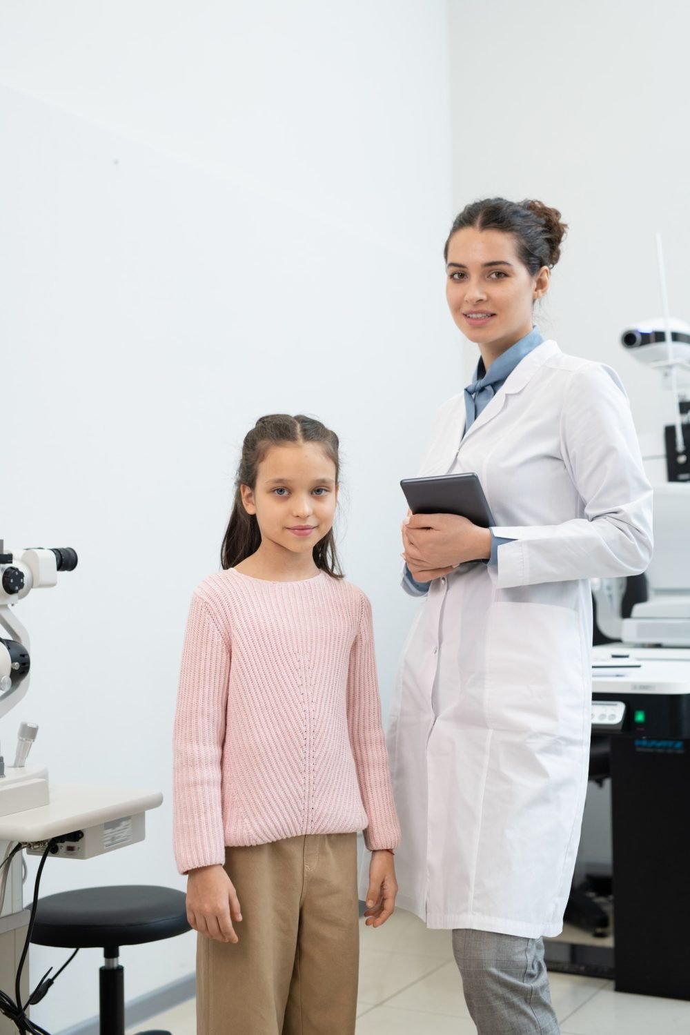 happy young female ophthalmologist in whitecoat with tablet and schoolgirl e1648178183881.jpg